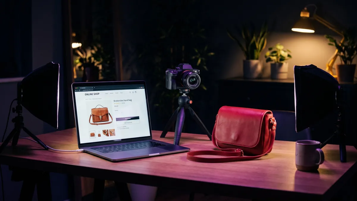 A laptop showing an online shop product page beside a mirrorless camera on a tripod, a red leather crossbody bag and a mug on a desk lit in purple